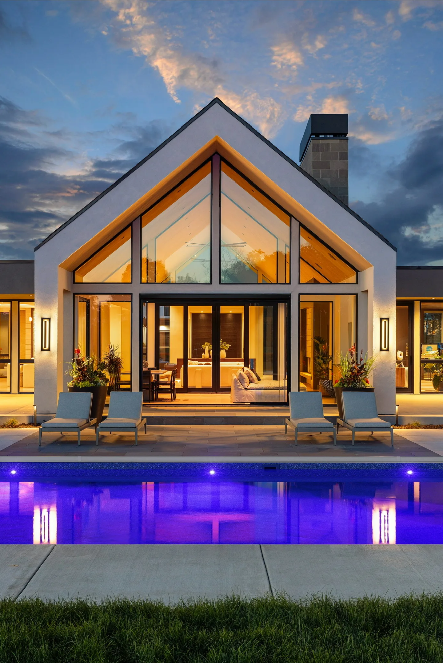 Modern gable home at dusk, glass walls glowing warm above a lit pool