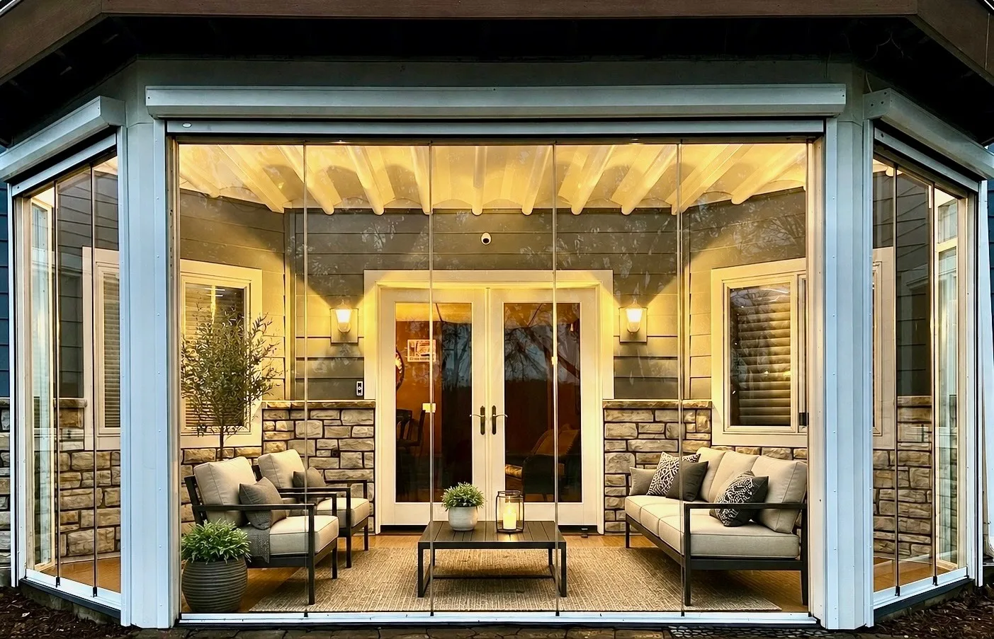 The same porch enclosed in frameless glass, warmly lit with seating inside